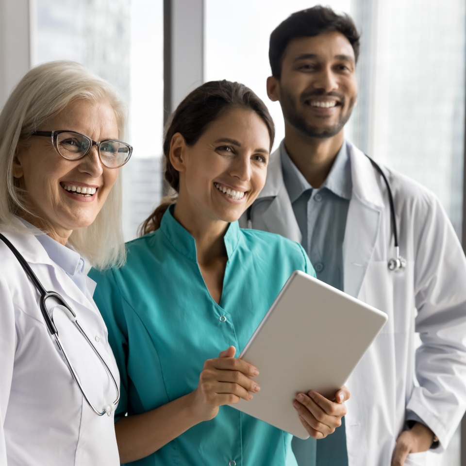 Smiling successful multiracial doctors friends stand at office corridor use digital tablet in teamwork look aside with smiles. Motivated modern clinic workers enjoy using modern tech for health care