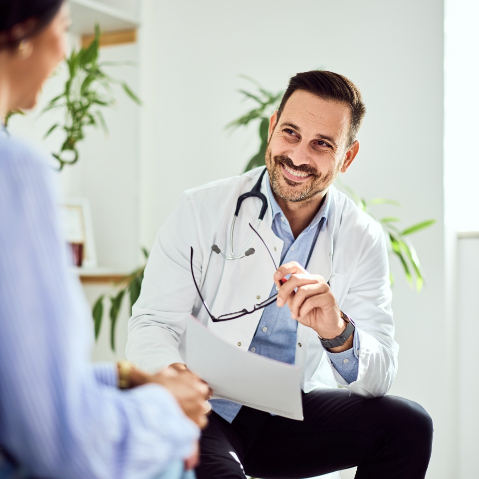 A physician attentively listens to a patient while holding their glasses, exuding professionalism and care. The well-lit setting and warm interaction highlight trust and effective communication in a medical consultation.