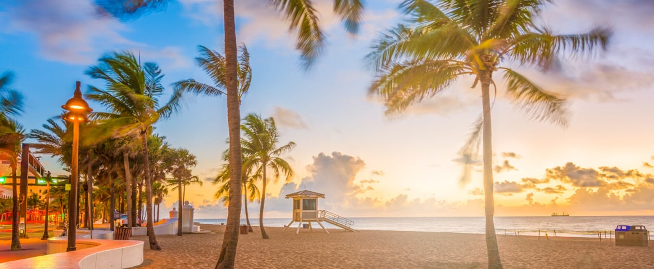 Palm trees and a lifeguard hut stand on a sandy beach at sunrise, with calm ocean waves and a partly cloudy sky in the background.
