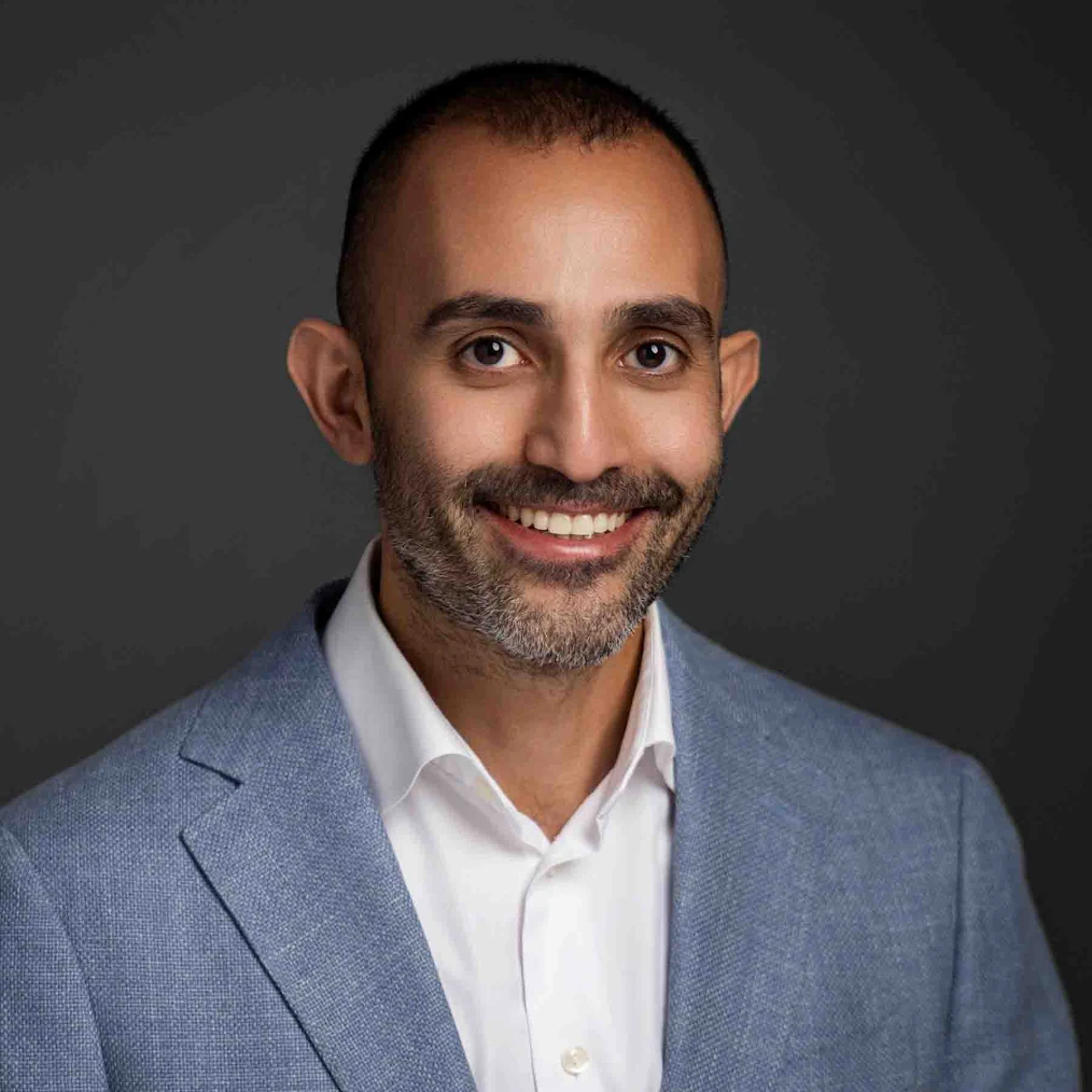A man with short hair and a beard, wearing a light blue suit jacket and white shirt, smiles in front of a dark gray background.