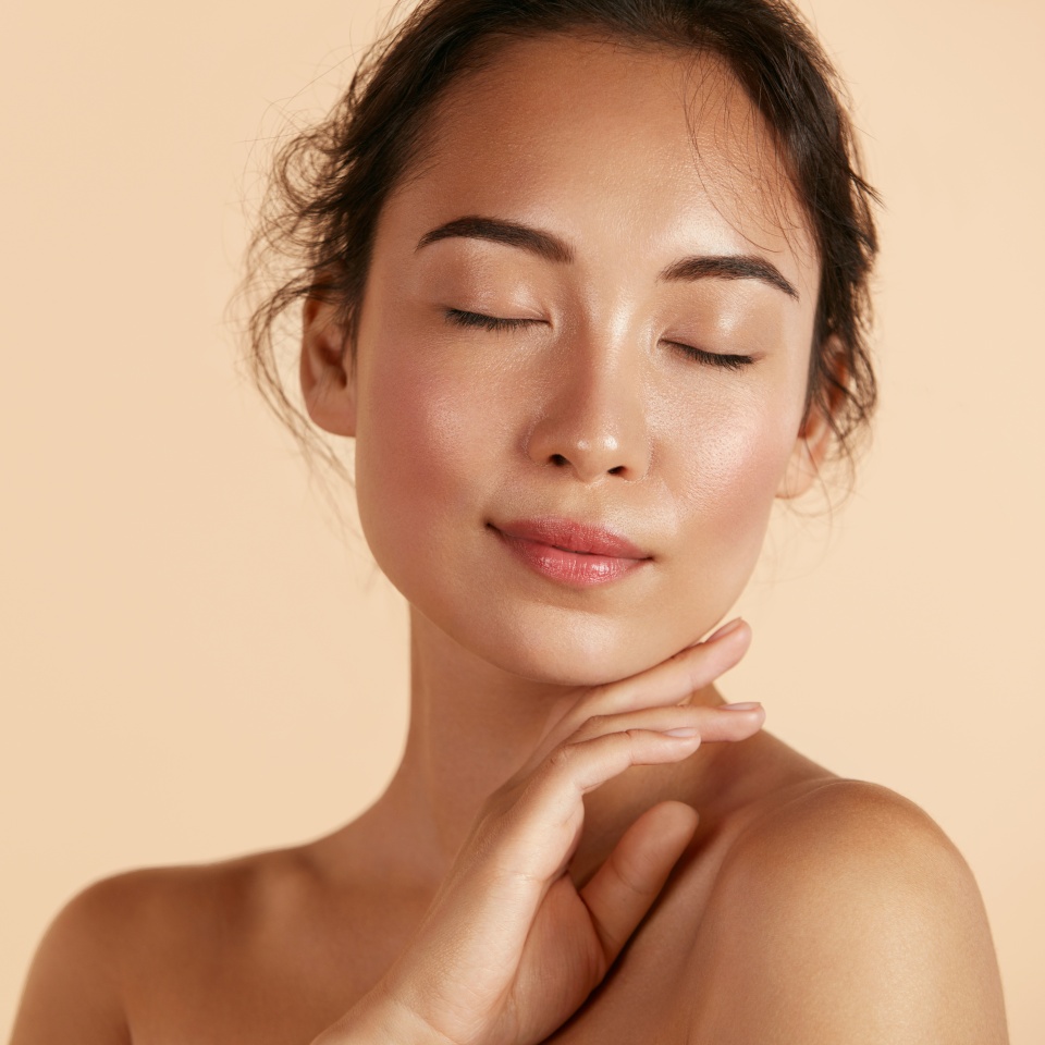 Beauty face. Woman with natural makeup and healthy skin portrait. Beautiful asian girl model touching fresh glowing hydrated facial skin on beige background closeup. Skin care concept