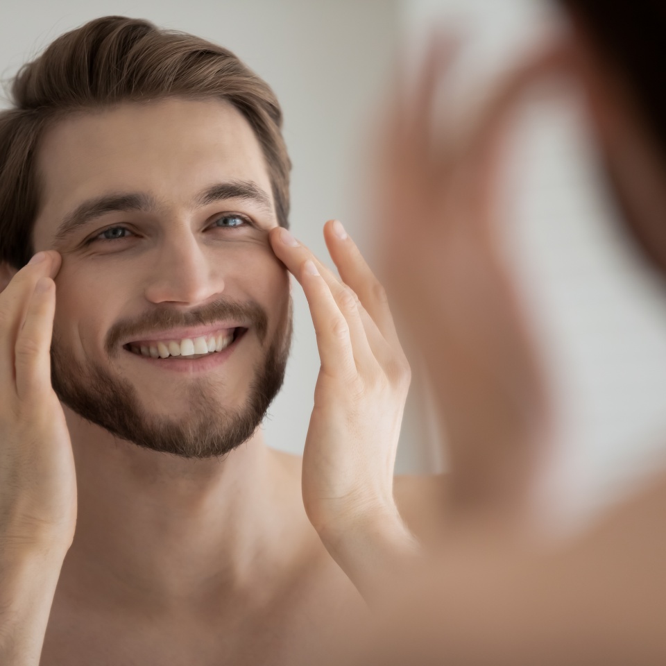 Close up mirror reflection smiling satisfied young man touching skin under eyes, applying anti aging moisturizing cream or lotion, standing in bathroom, enjoying skincare procedure, morning routine