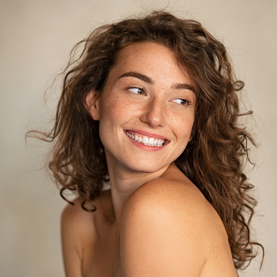 Portrait of smiling girl enjoying beauty treatment on beige background. Beautiful natural woman looking at copy space, spa and wellness concept. Carefree laughing woman with bare shoulders isolated.