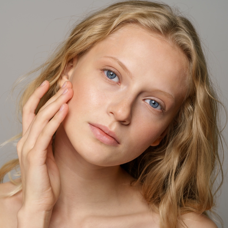 Closeup of pretty young woman face with blue eyes, curly natural blonde hair, has no makeup, touching her soft skin, standing shirtless with bare shoulders, looking at camera. Studio grey background