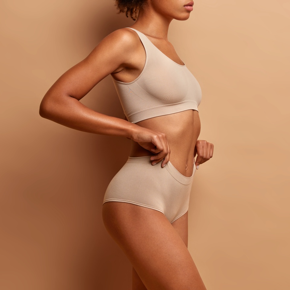 Profile view of slim dark skinned woman dressed in cropped top and panties demonstrates her perfect body slender legs poses against brown background