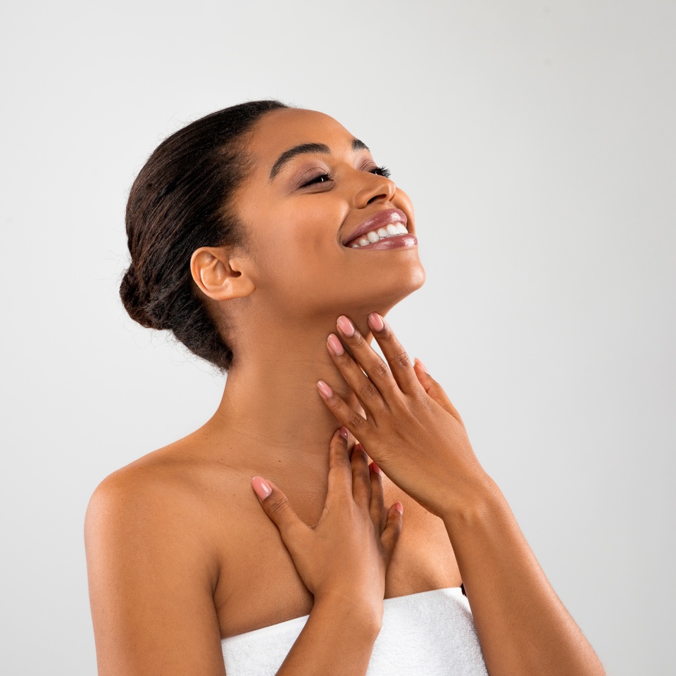 Essential Beauty. Joyful young black woman wrapped in towel touching her soft flawless skin on neck, attractive african american lady enjoying result of beauty treatments, grey background, copy space