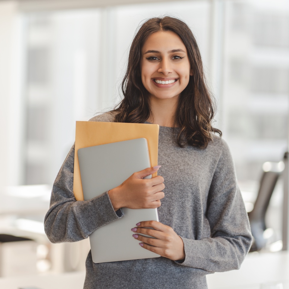 Smiling confident Indian woman wearing casual clothes holding laptop and documents, looking at camera at modern office. Good looking businesswoman posing for picture. Concept of successful business