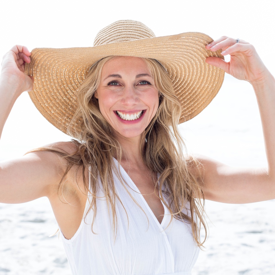 Smiling blonde in white dress looking at camera and wearing straw hat at the beach