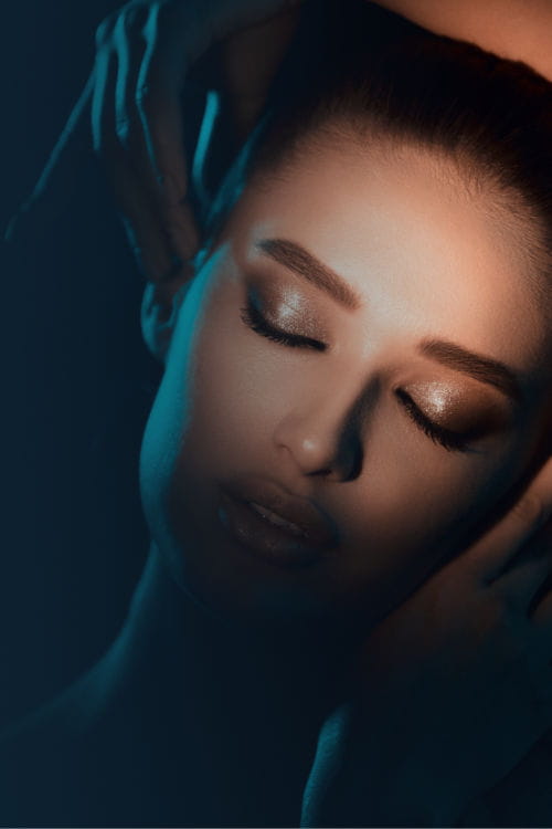 A woman with closed eyes and shimmering eye makeup is softly illuminated by a narrow beam of light, with hands gently touching her head and face.